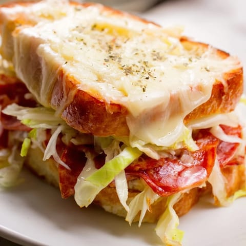 Two halves of a toasted Italian Sub Grilled Cheese reveal stacked salami and pepperoni beside crisp kettle chips and fresh tomato salad.